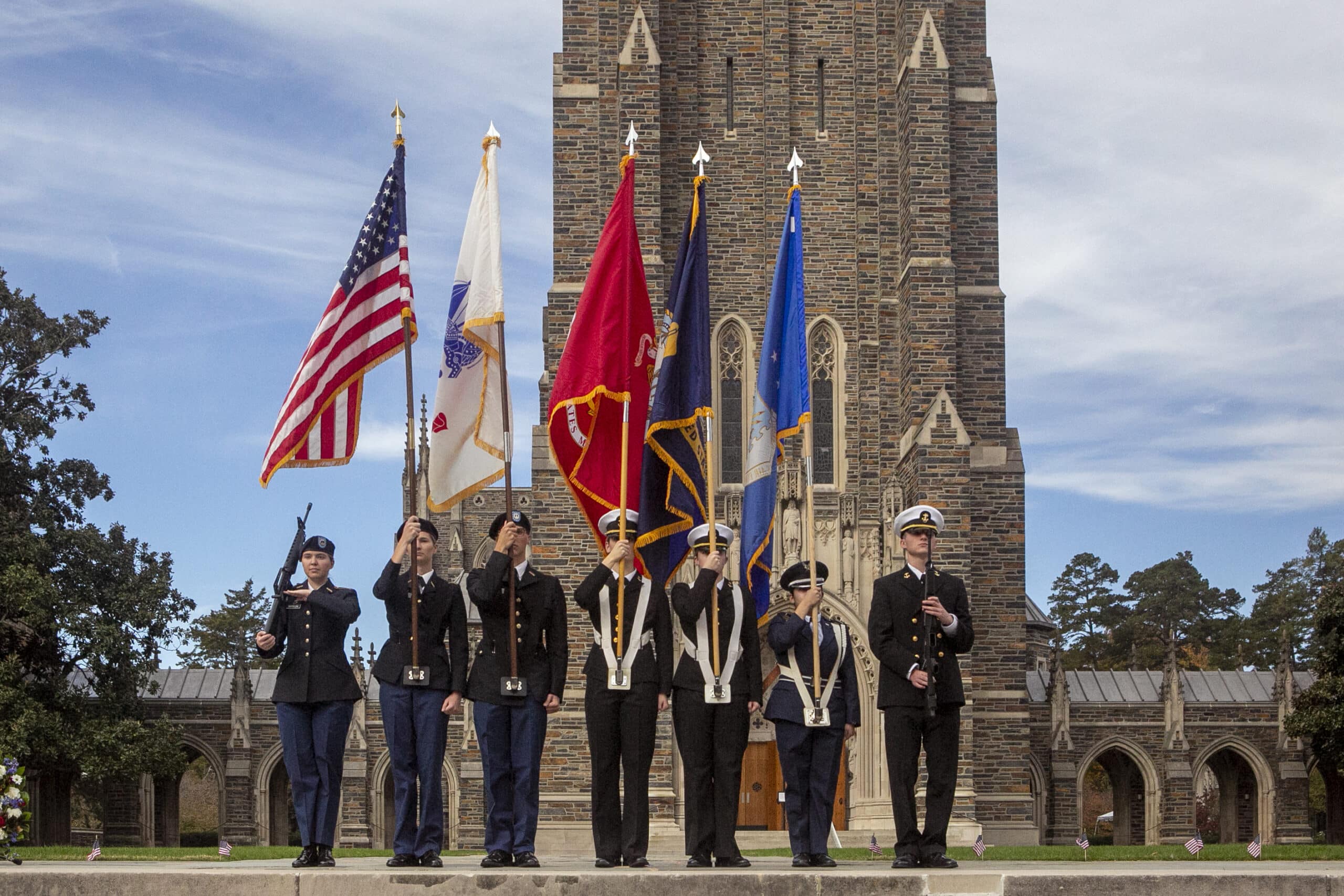 a uniformed colorguard carries flags in front of a Collegiate Gothic building